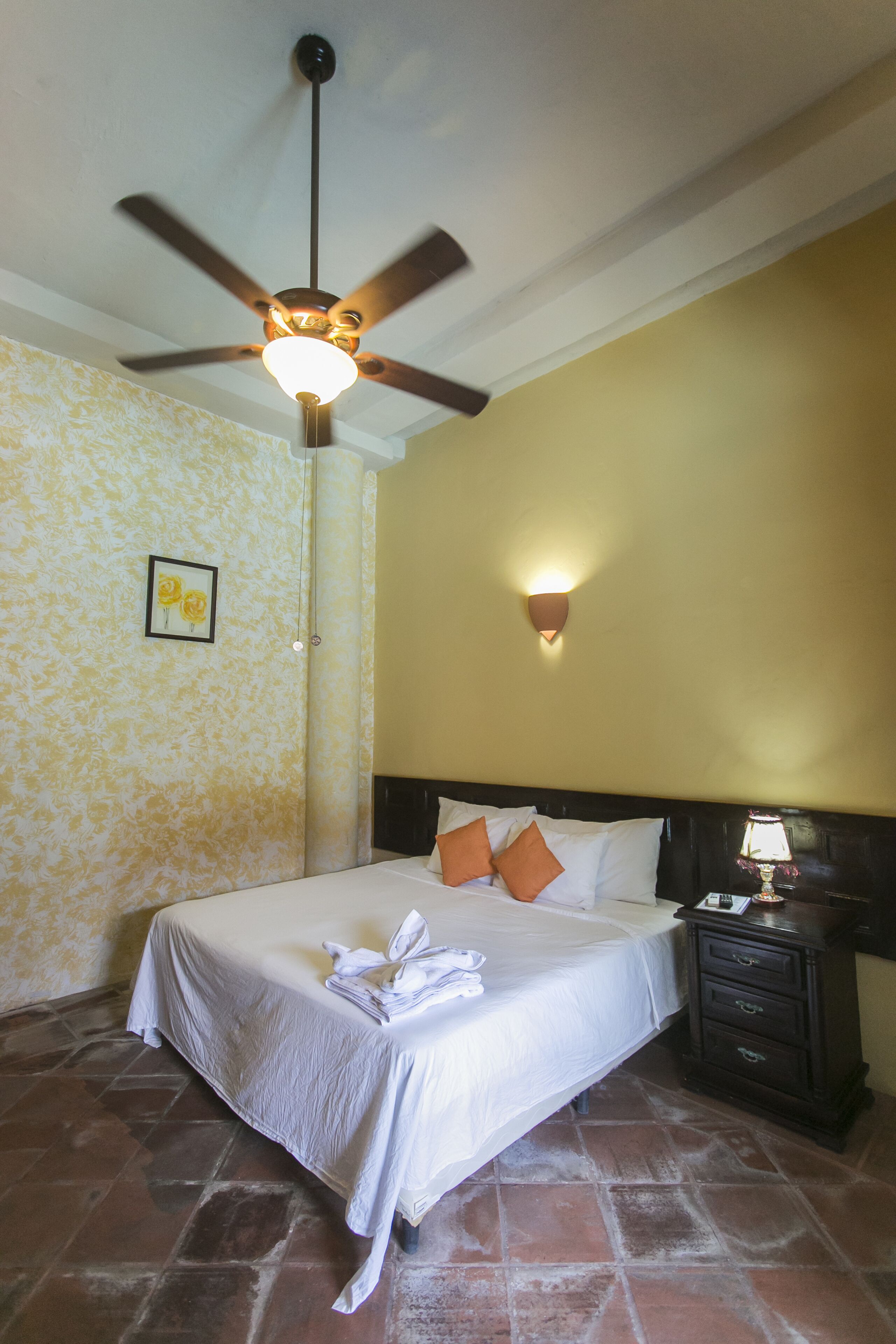 A hotel room with a bed, a ceiling fan, a nightstand, and a wall-mounted light.