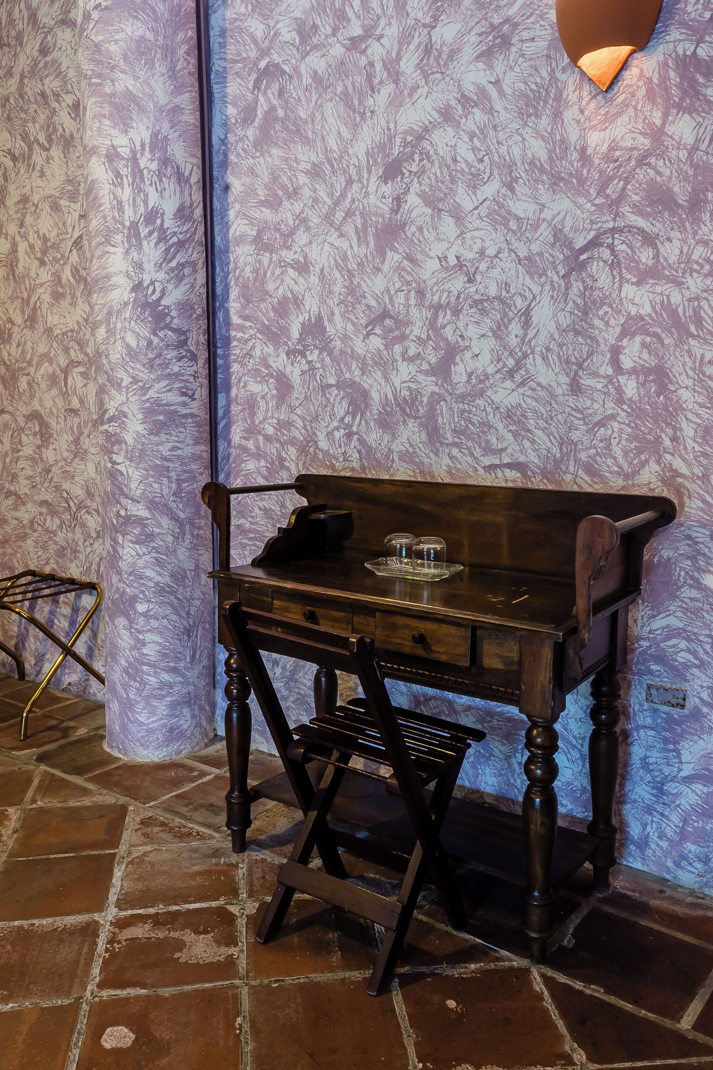 A wooden desk with drawers and a chair, a small table with a glass, and a wall with a textured pattern.