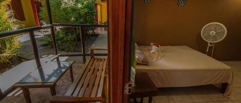 A room with a bed, a small table, a bench, and a fan. There is a view of a garden outside.
