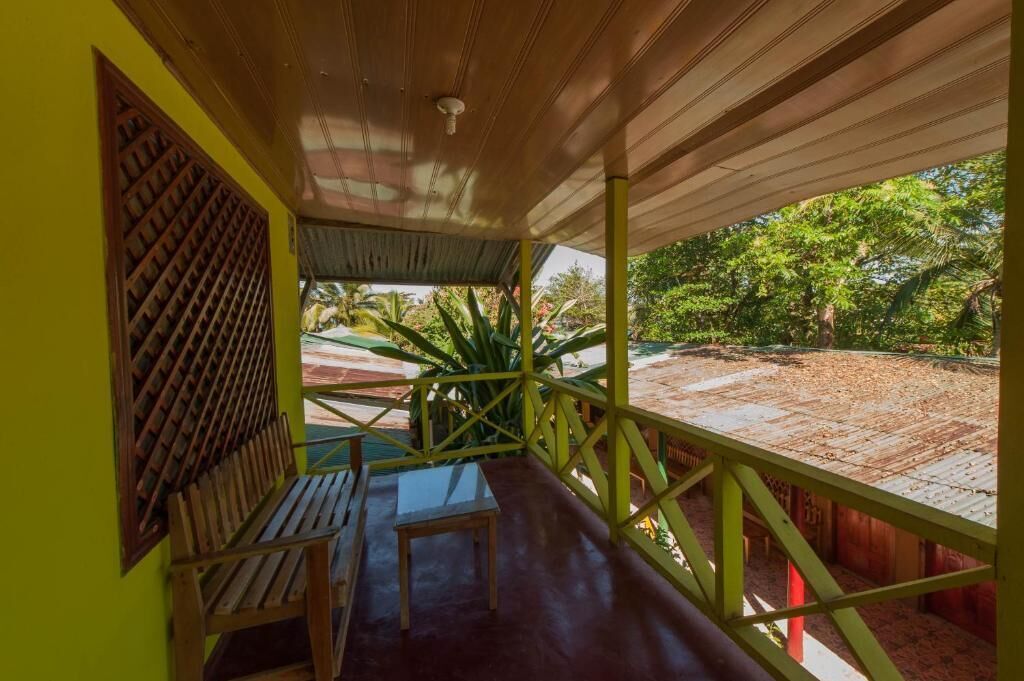 A covered porch with a wooden bench and a small table, overlooking a garden with large plants and a red-roofed structure.