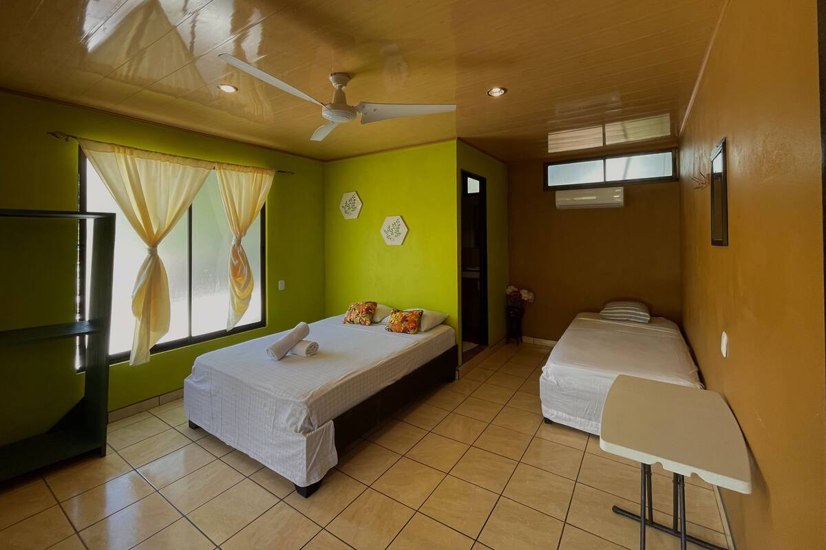 A room with two beds, a ceiling fan, a window with curtains, and a small table.