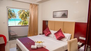 A hotel room with a bed, a desk, a chair, a window with a view of palm trees and the ocean, and a framed picture on the wall.