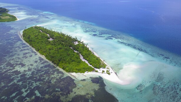 Vista aérea de una isla tropical con una playa estrecha, rodeada de aguas azules cristalinas y exuberante vegetación.