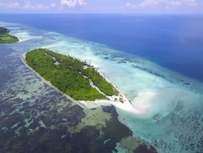 Vista aérea de uma ilha tropical com uma praia estreita, cercada por águas azul-claras e vegetação exuberante.