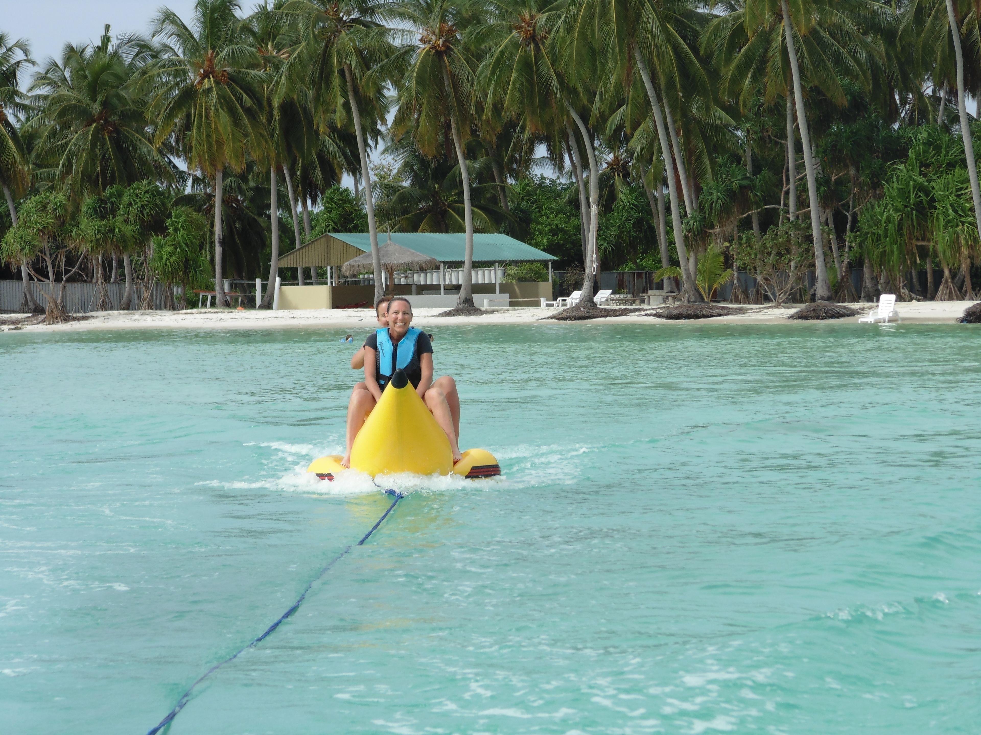 Une personne conduisant un véhicule nautique jaune sur une eau turquoise et limpide, entourée de palmiers et d’une plage avec des chaises longues.