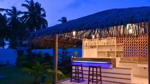 Zona de bar con techo de paja, taburetes de madera y mostrador con estantes llenos de botellas.