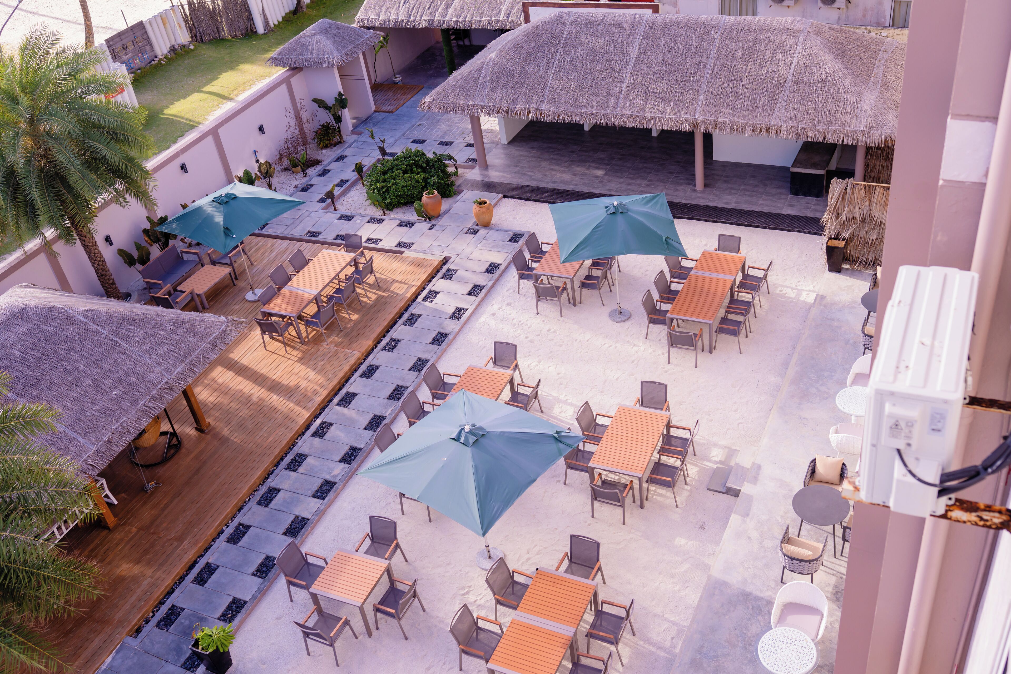 An outdoor patio with multiple tables and chairs, surrounded by thatched umbrellas, and a pool area.