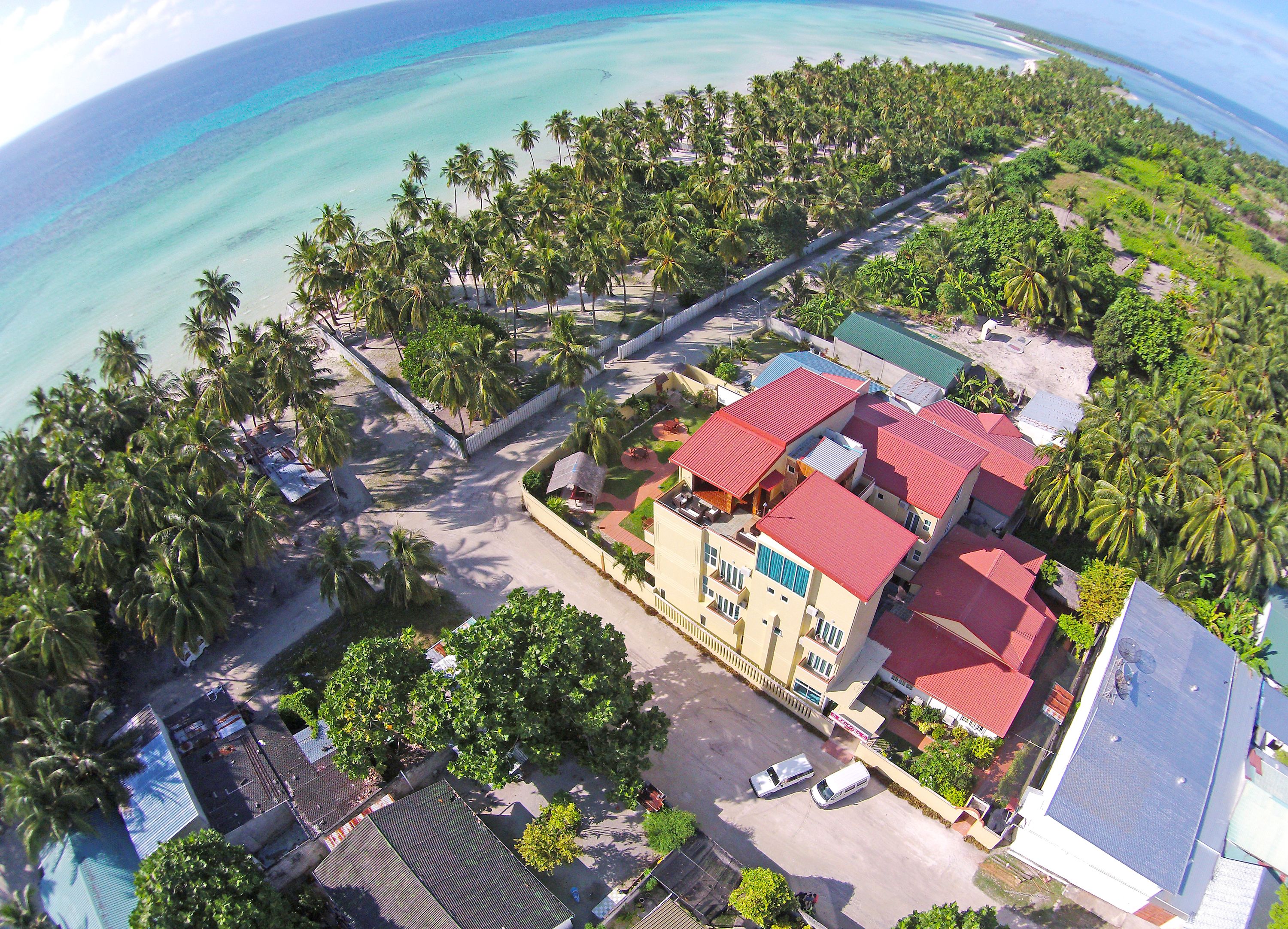 An aerial view of a coastal area with a hotel featuring a red-roofed building, surrounded by palm trees and a beach.