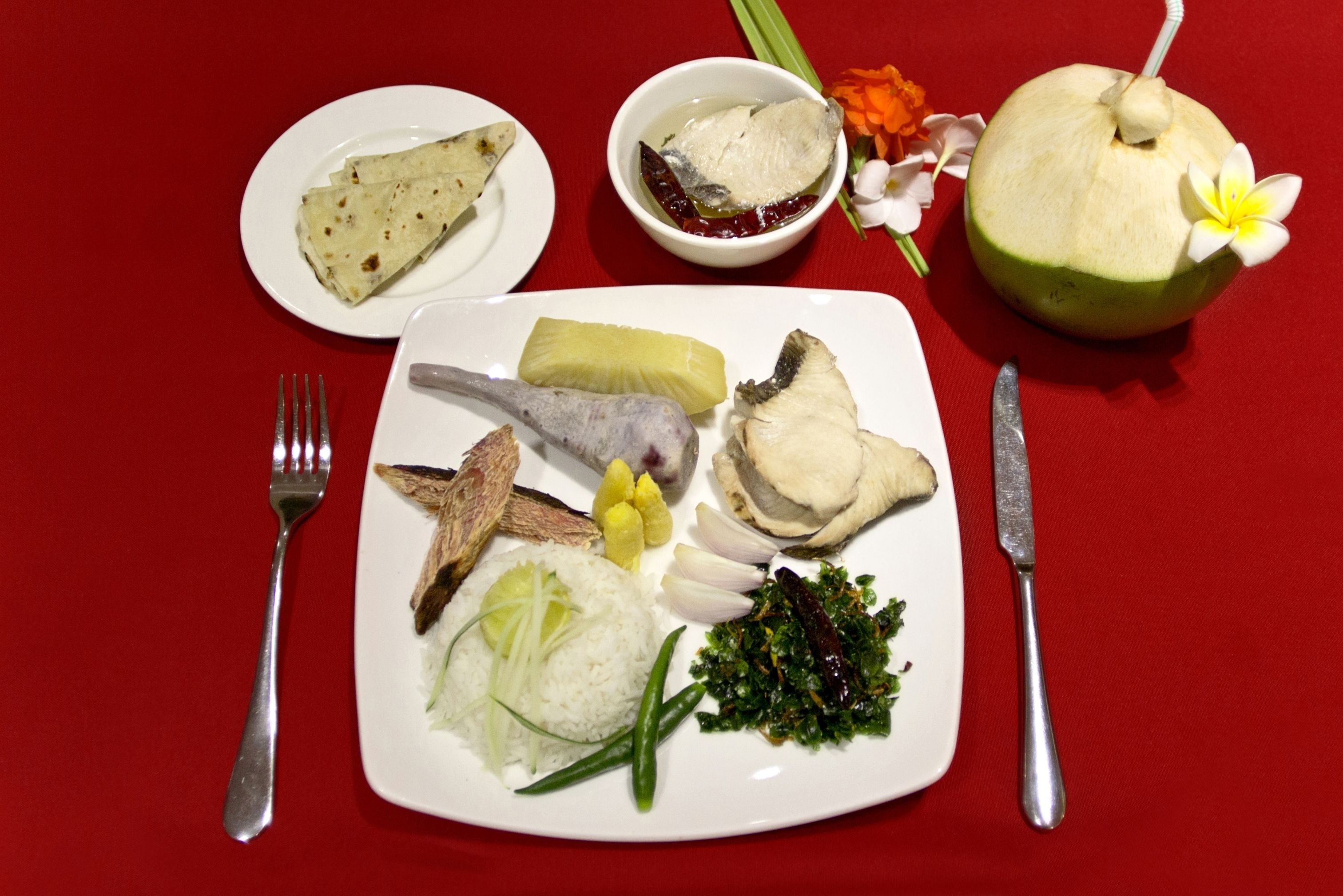 A plate with various dishes including fish, rice, and vegetables, accompanied by a coconut drink and a small bowl of sauce.