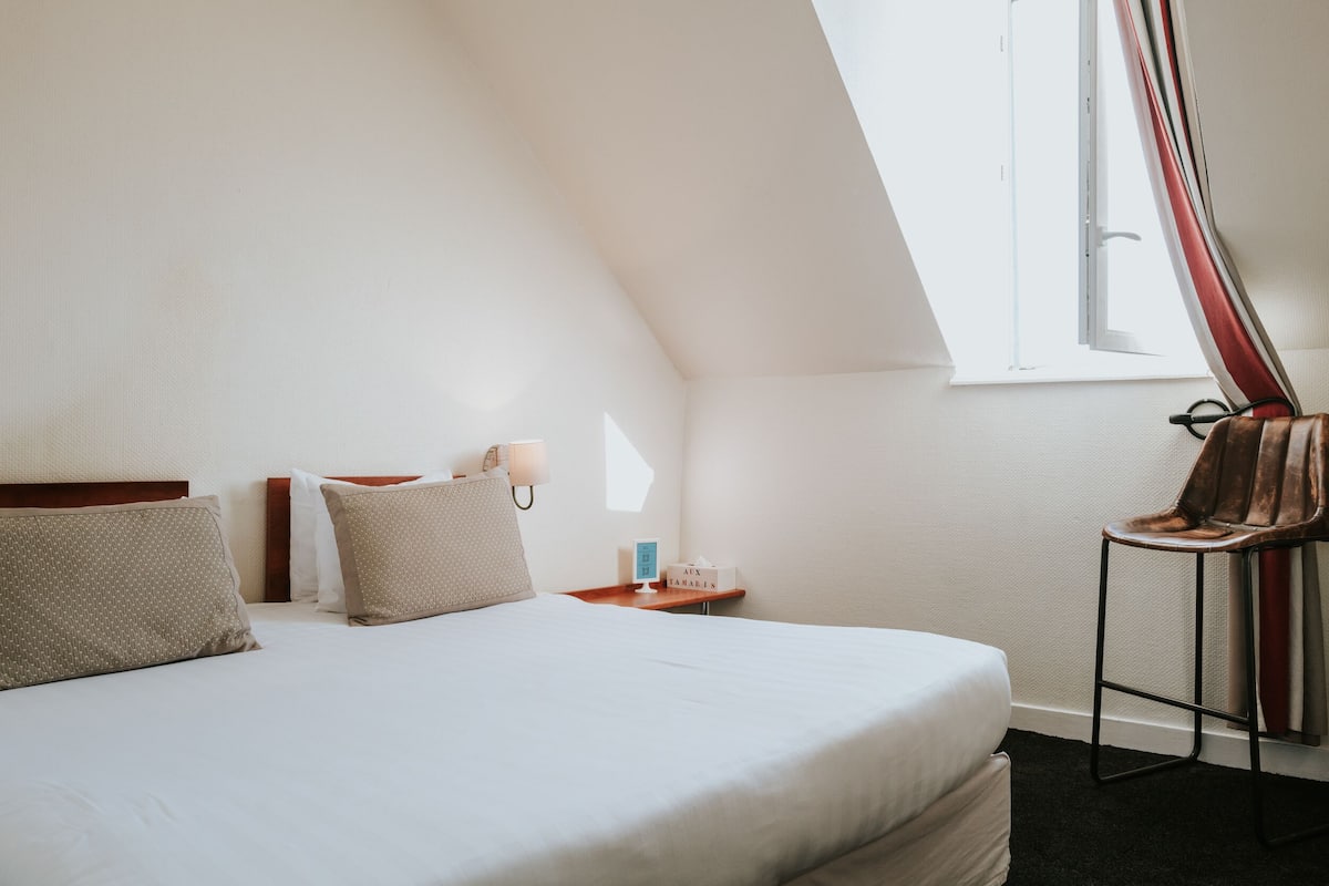 Ein Hotelzimmer mit einem großen Bett, einem Nachttisch, einem Sessel und einem Fenster mit Vorhängen.