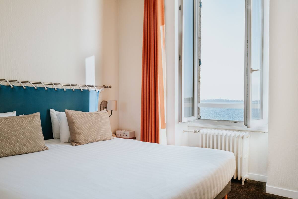 Ein Hotelzimmer mit Bett, Nachttisch, Lampe, einem Fenster mit Vorhängen und Blick aufs Meer.
