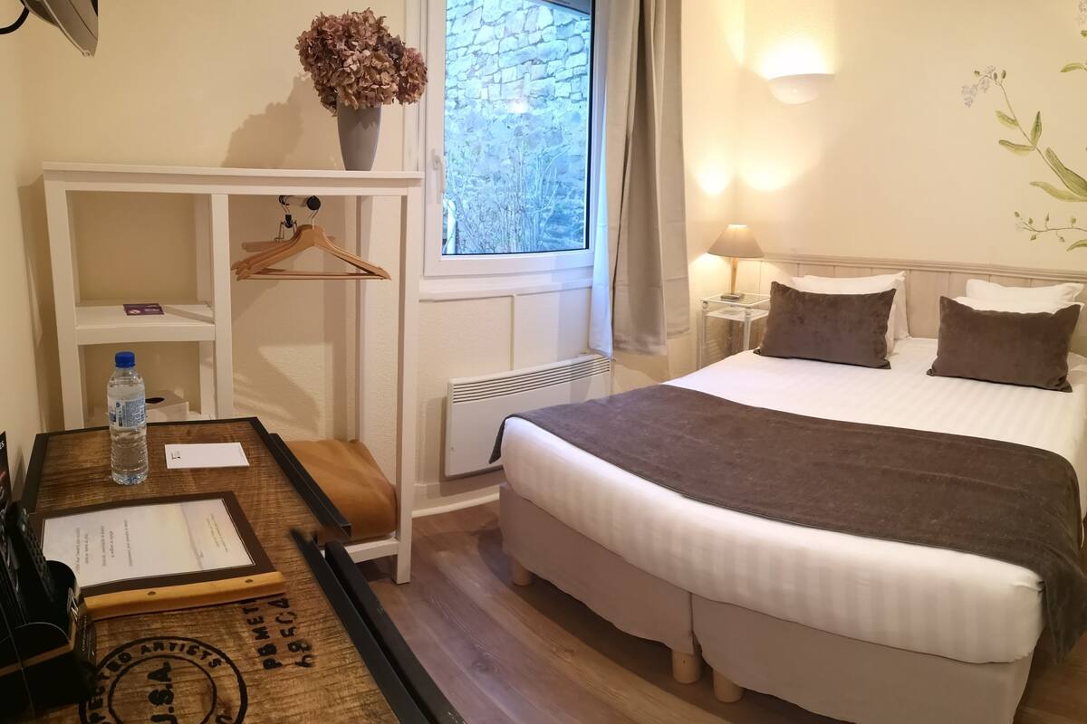 Ein ordentlich eingerichtetes Hotelzimmer mit einem großen Bett, Nachttischen, einem Kleiderhaken und einem Fenster mit Blick auf eine Steinmauer. Auf einem Regal steht eine Vase mit getrockneten Blumen.