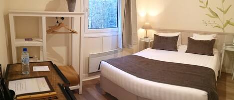 A neatly arranged hotel room with a large bed, bedside tables, a hanging coat rack, a window with a view of a stone wall, and a vase of dried flowers on a shelf.