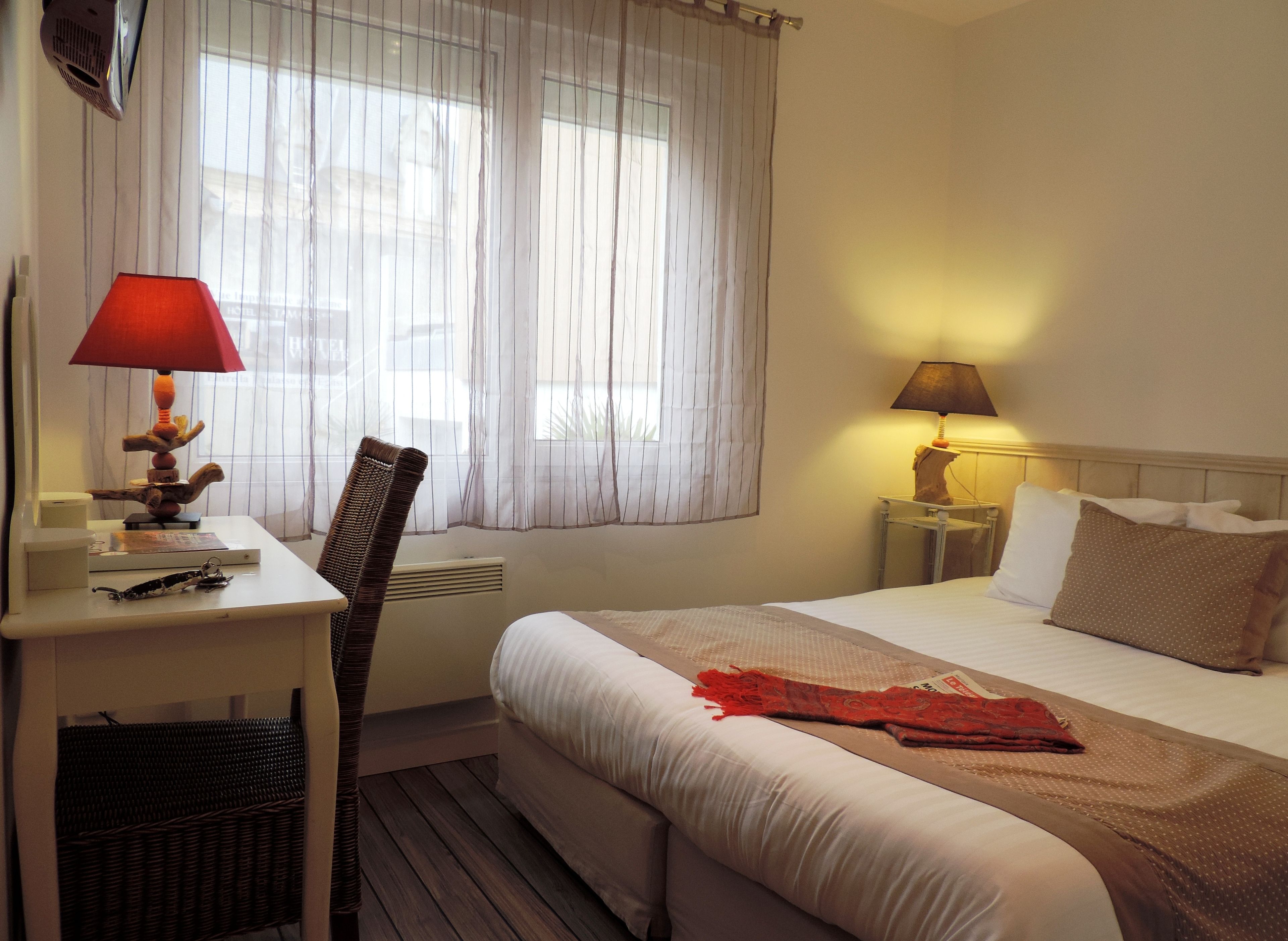 Une chambre d’hôtel avec un lit, un bureau, une chaise, une lampe et une fenêtre avec des rideaux.