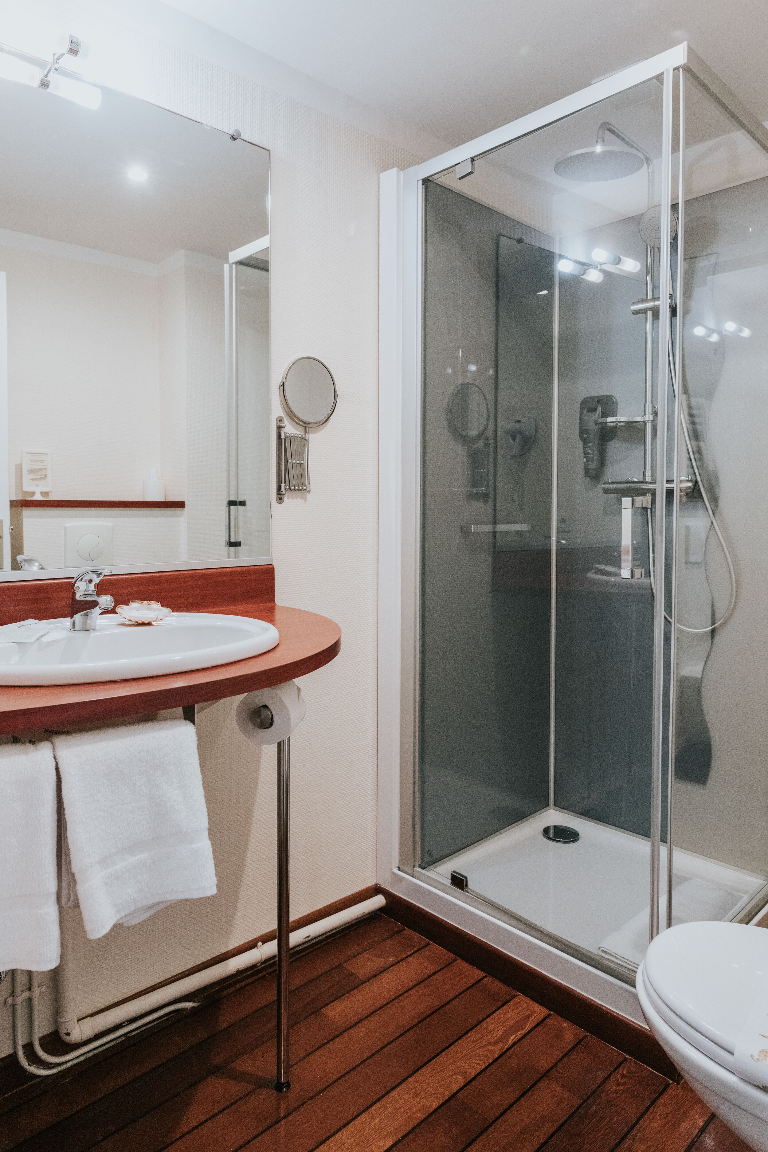 Un baño con piso de madera, un lavamanos blanco con espejo y una ducha con mampara de vidrio.