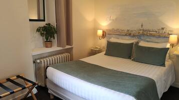 A hotel room with a bed, a nightstand, a lamp, a plant, and a painting on the wall.