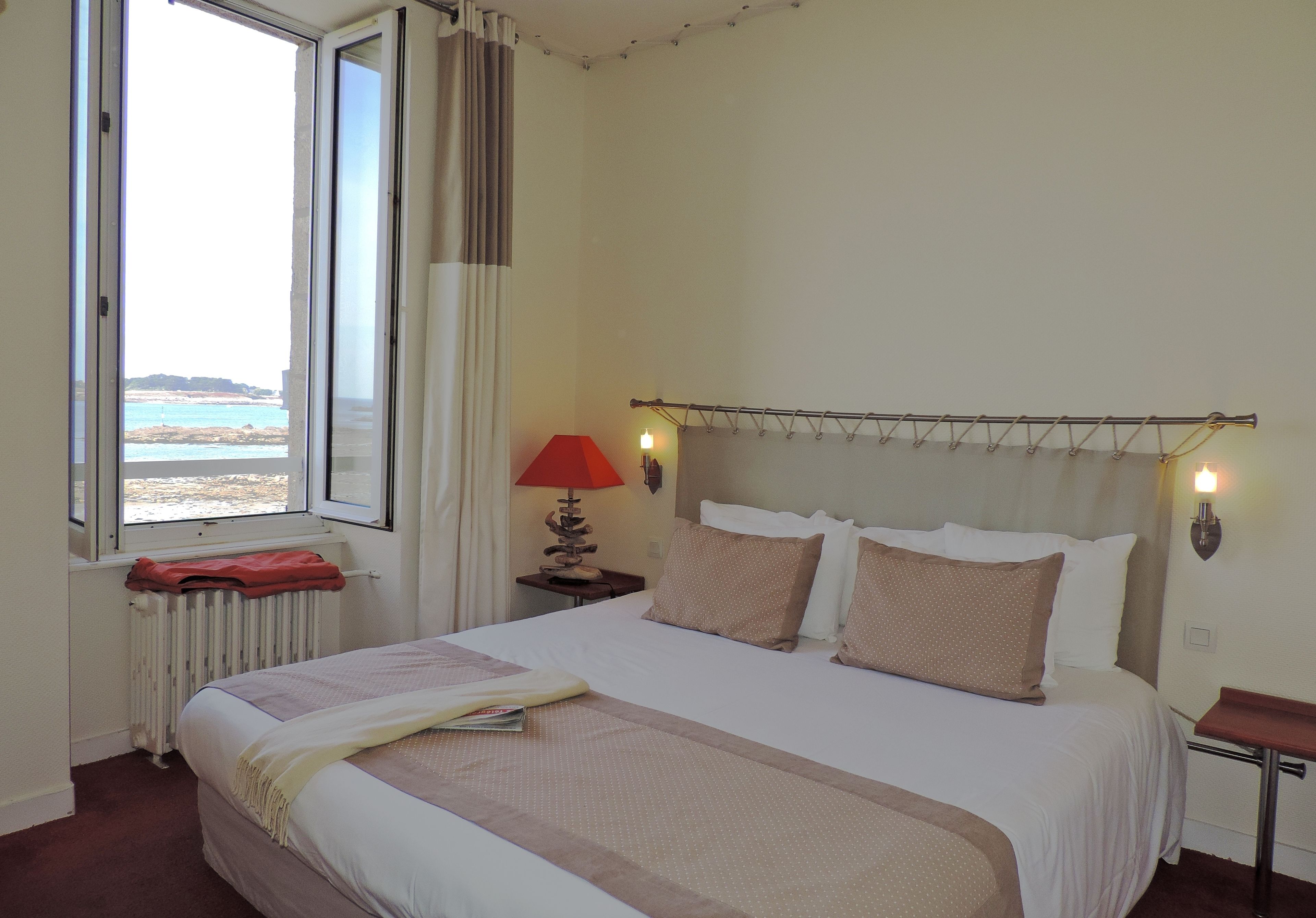 A hotel room with a large bed, a window with a view of the beach, a bedside table with a lamp, and a red blanket on the radiator.