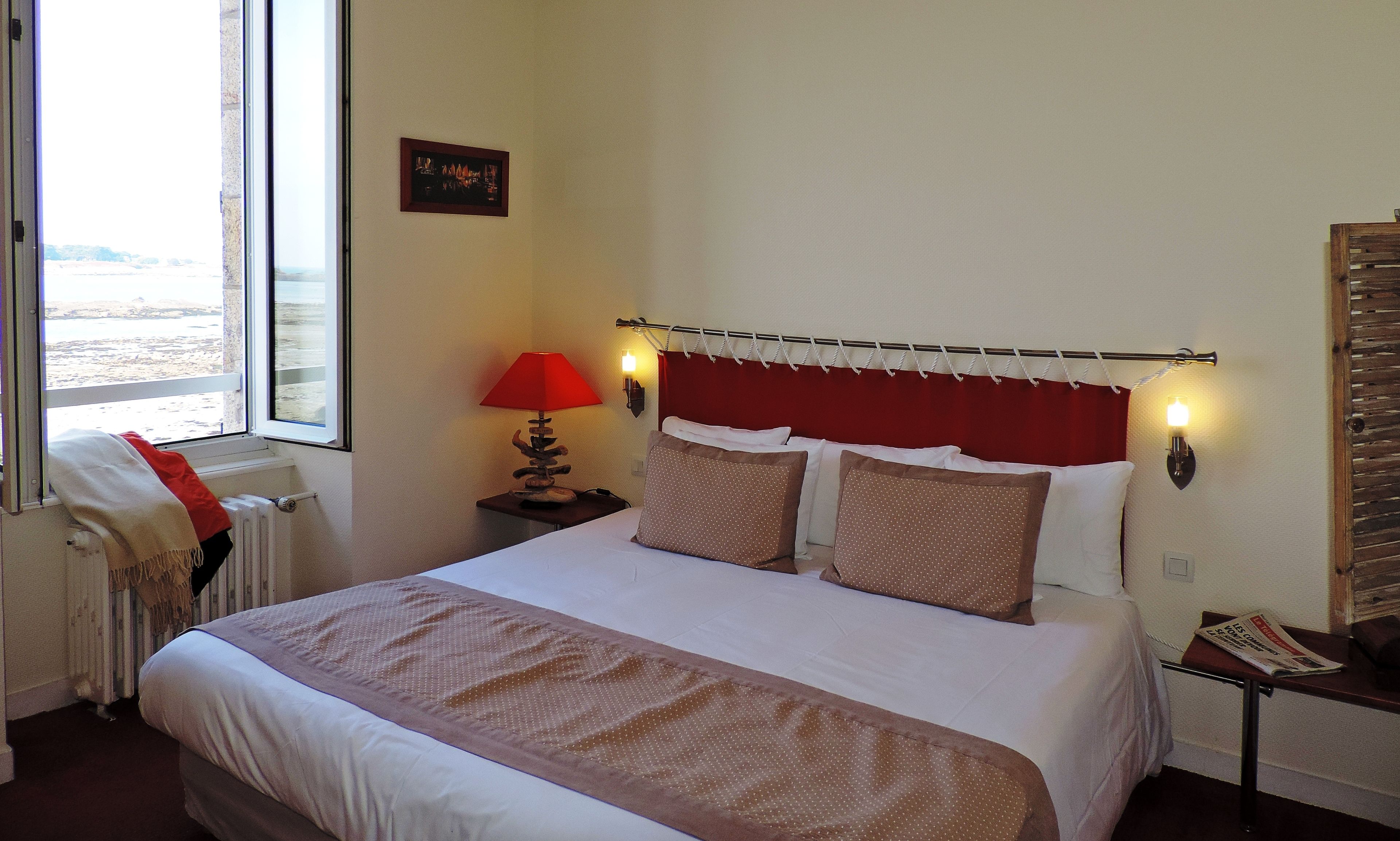 A hotel room with a bed, a window with a view, a lamp, and a small table with a newspaper.