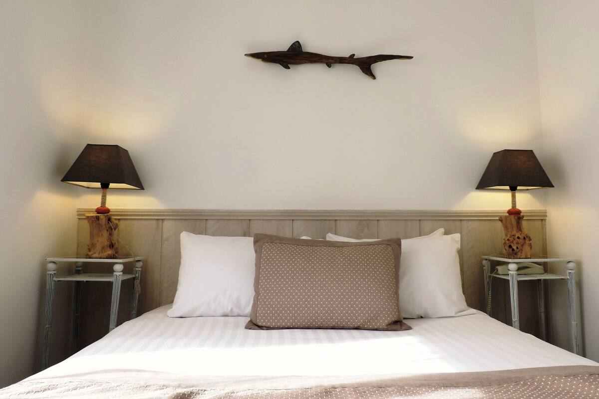 Ein ordentlich gemachtes Bett mit weißen und gemusterten Kissen, zwei Nachttischlampen und einer Wanddekoration mit Fischmotiv.