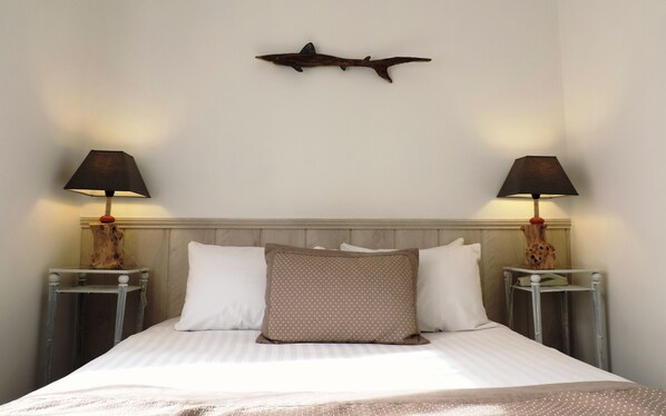 Una cama bien hecha con almohadas blancas y estampadas, dos lámparas de noche y una decoración de peces colgada en la pared.