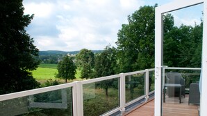 A balcony with a wooden deck, glass railings, and a view of a green landscape with trees and hills. - Hotel Restaurant Vijlerhof (Vijlen)