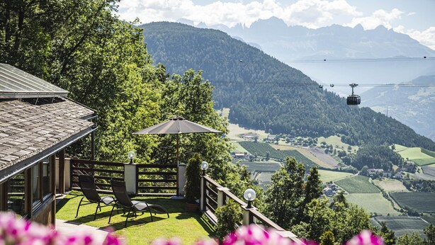 En terrasse med udsigt over et bjerglandskab, en kabelbane i det fjerne, samt en stol og parasol.