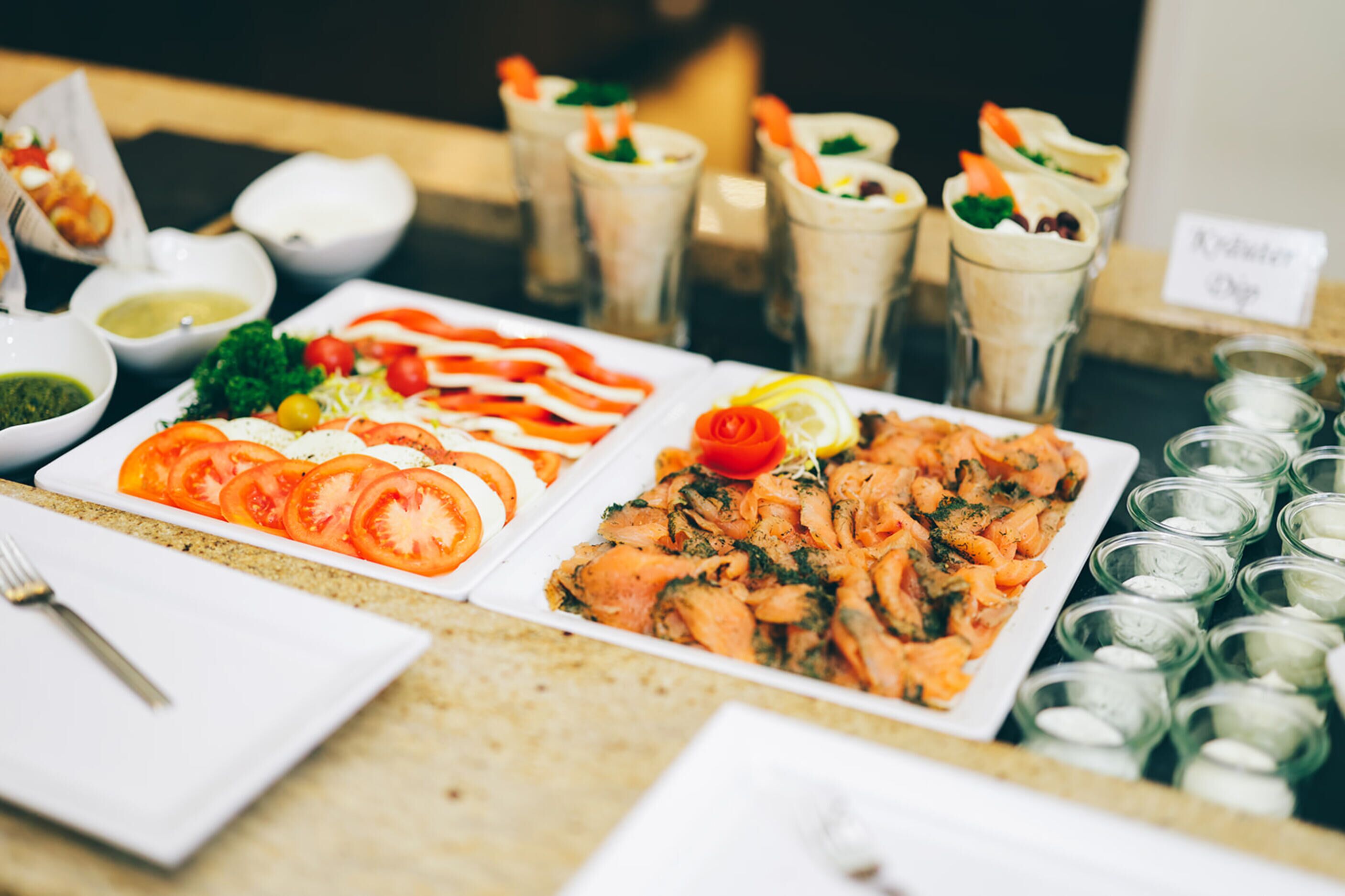 Buffet com diversos pratos, incluindo tomates fatiados, salmão e vários molhos.