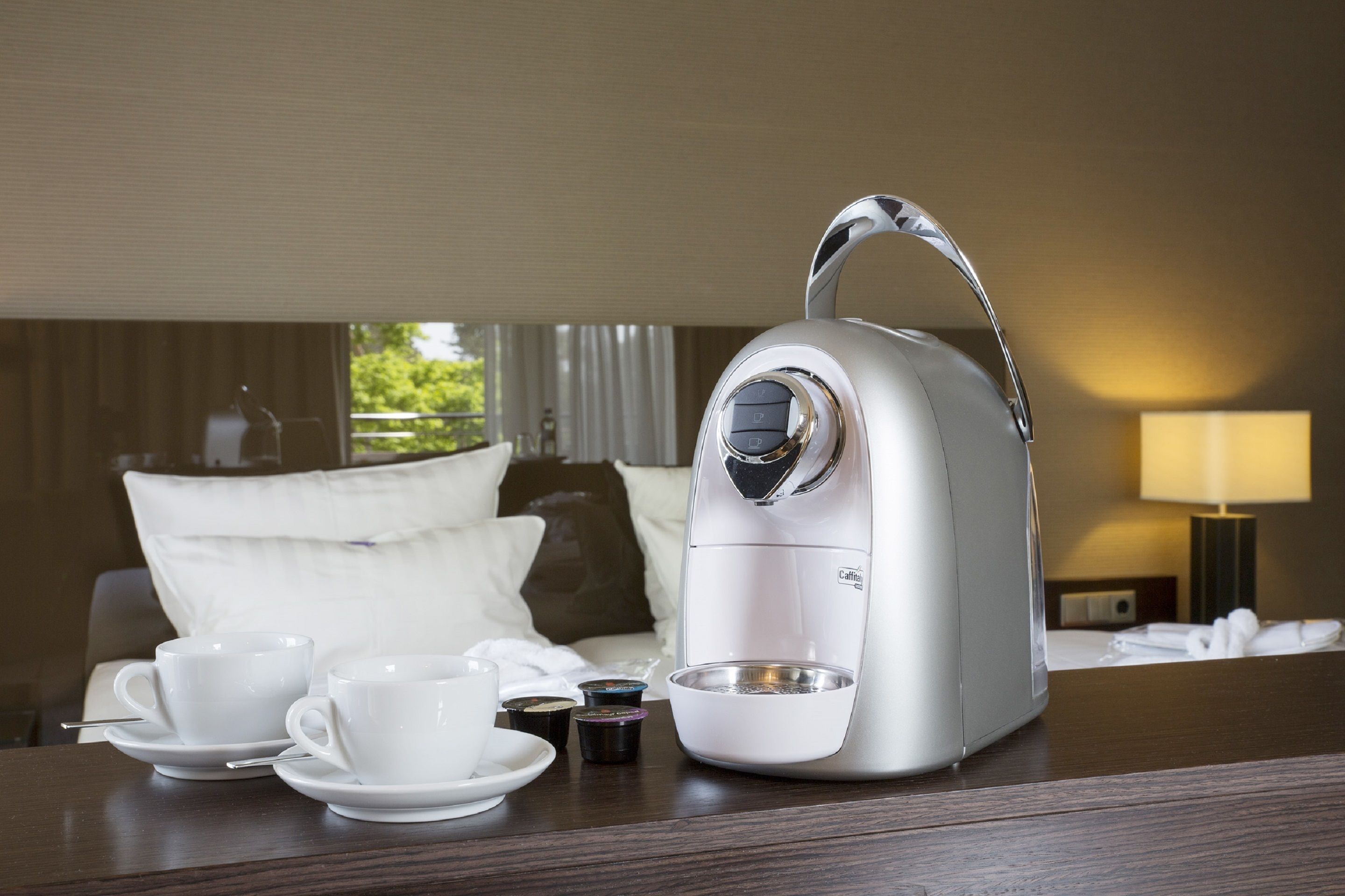 Eine Kaffeemaschine auf einem Tisch mit zwei weißen Tassen und Untertassen, einer kleinen Schale und einer Kaffeekapsel-Auswahl.