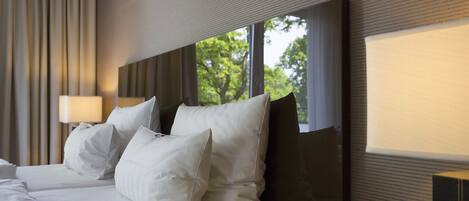 Habitación de hotel con una cama grande, varias almohadas, una lámpara de noche y una ventana con vistas a árboles.