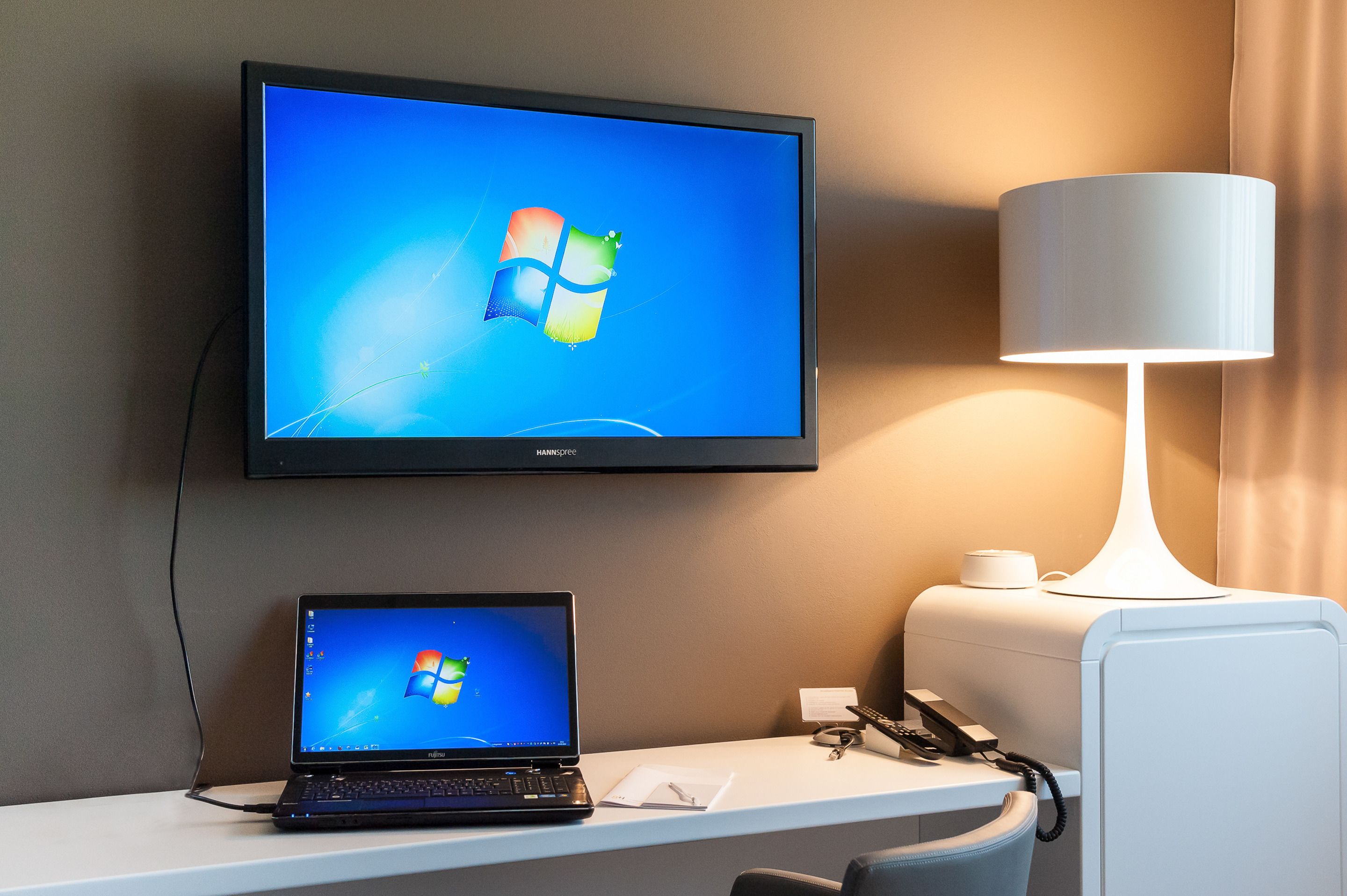 A hotel room with a flat-screen TV displaying the Windows logo, a laptop on a desk, a lamp, and a telephone.