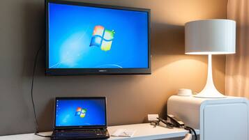 Ein Hotelzimmer mit Flachbildfernseher, der das Windows-Logo anzeigt, einem Laptop auf einem Schreibtisch, einer Lampe und einem Telefon.
