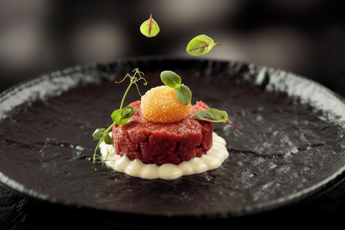A dish with a spherical garnish on top, surrounded by greens, on a dark plate.