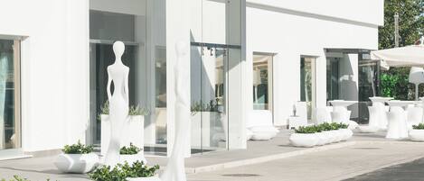 Un edificio moderno con fachada blanca, amplias ventanas y zonas de estar al aire libre con macetas y esculturas blancas.