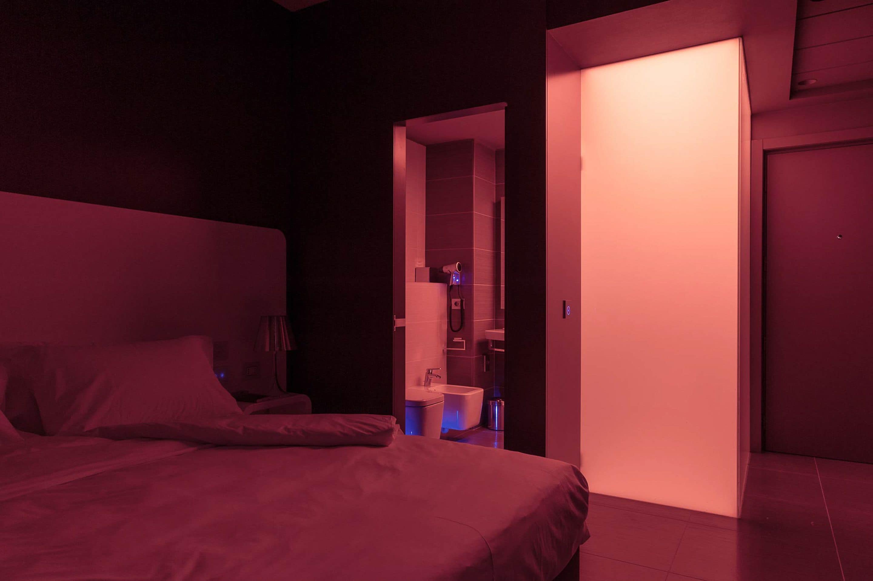 Une chambre d’hôtel faiblement éclairée, avec un lit, un petit bureau et une salle de bain visible à travers une porte entrouverte.