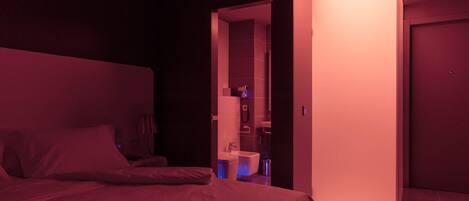 Une chambre d’hôtel faiblement éclairée, avec un lit, un petit bureau et une salle de bain visible à travers une porte entrouverte.