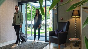 A modern hotel lobby with a glass entrance, a sofa, a lamp, and a magazine stand. - Parkhotel zur Linde (Silandro)