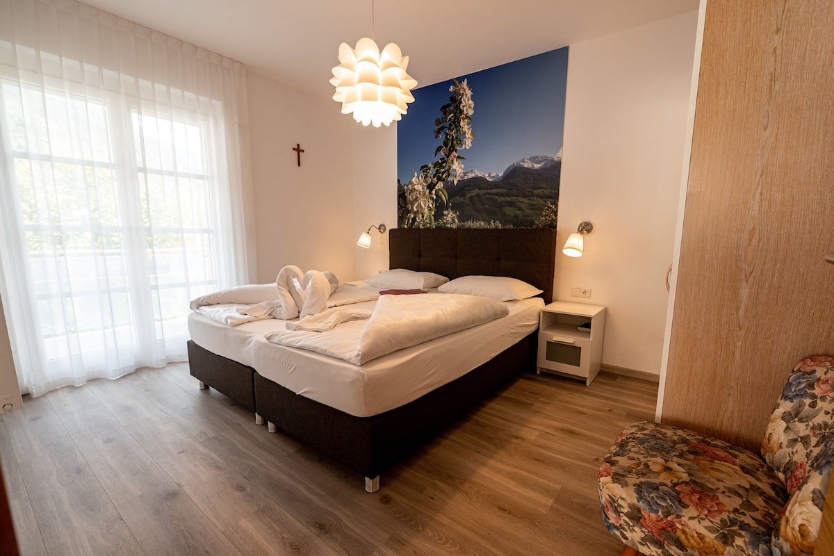 A bedroom with a bed, bedside lamps, a nightstand, a floral armchair, and a mountain landscape mural on the wall.