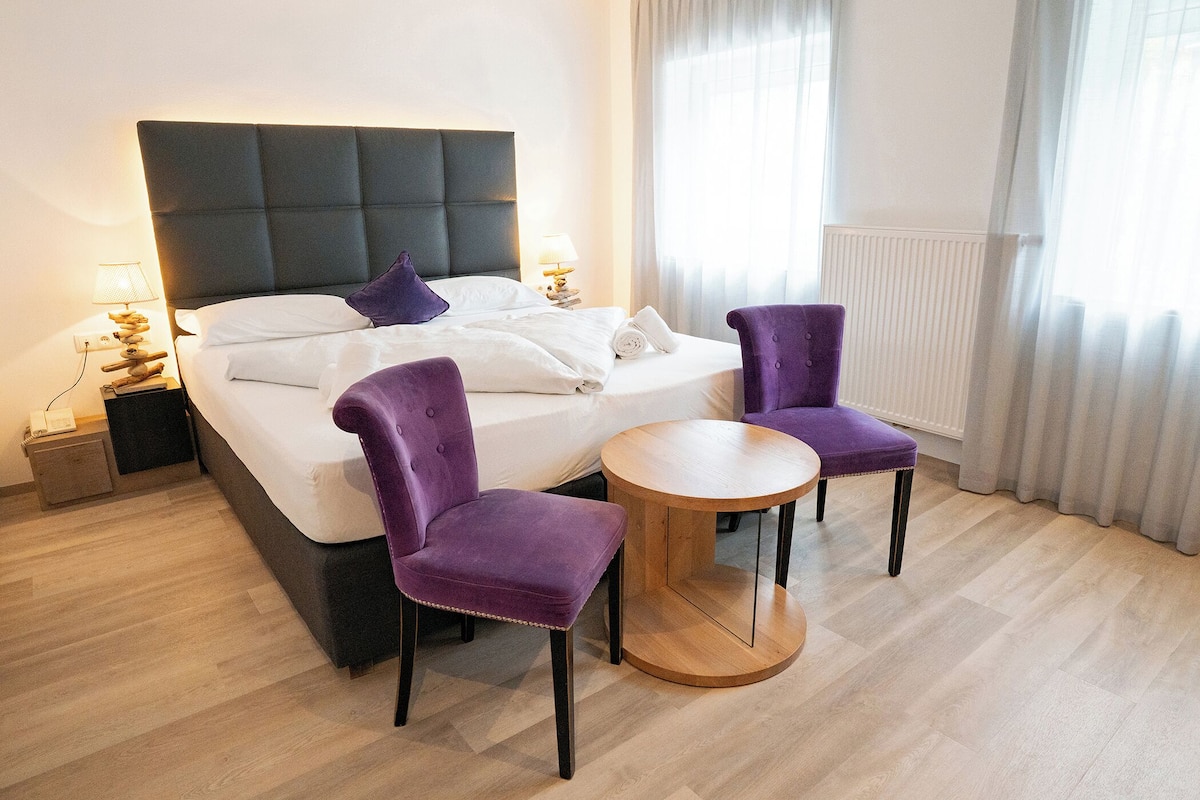 A hotel room with a bed, two purple chairs, a small wooden table, and a nightstand with a lamp.