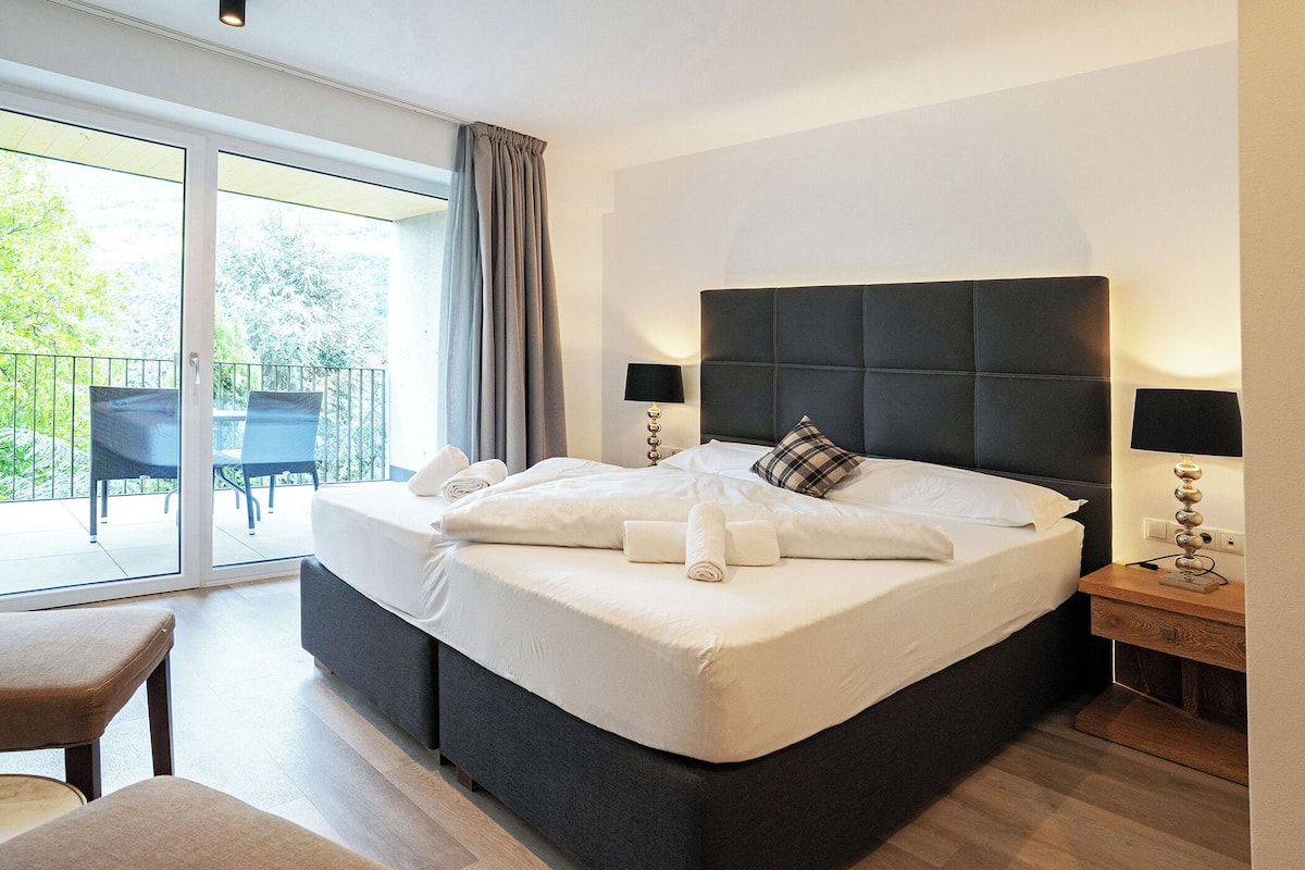 A hotel room with a large bed, a desk, and a chair. There is a balcony with chairs and a table.