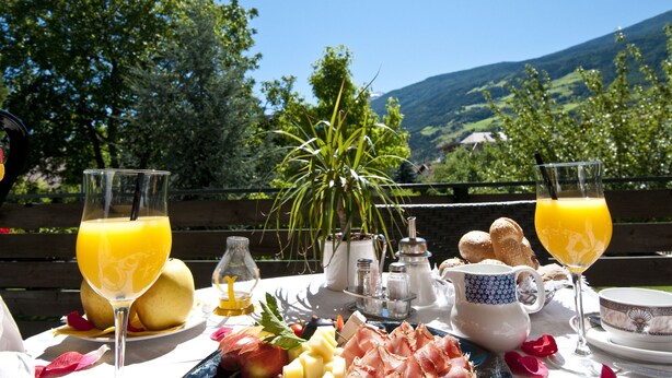 A table set with breakfast items including bread, fruit, and a jug of juice, with a scenic view in the background. - Parkhotel zur Linde (Silandro)