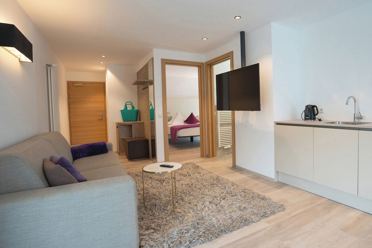 A modern living room with a sofa, a small round table, and a kitchen area with a sink and a flat-screen TV.