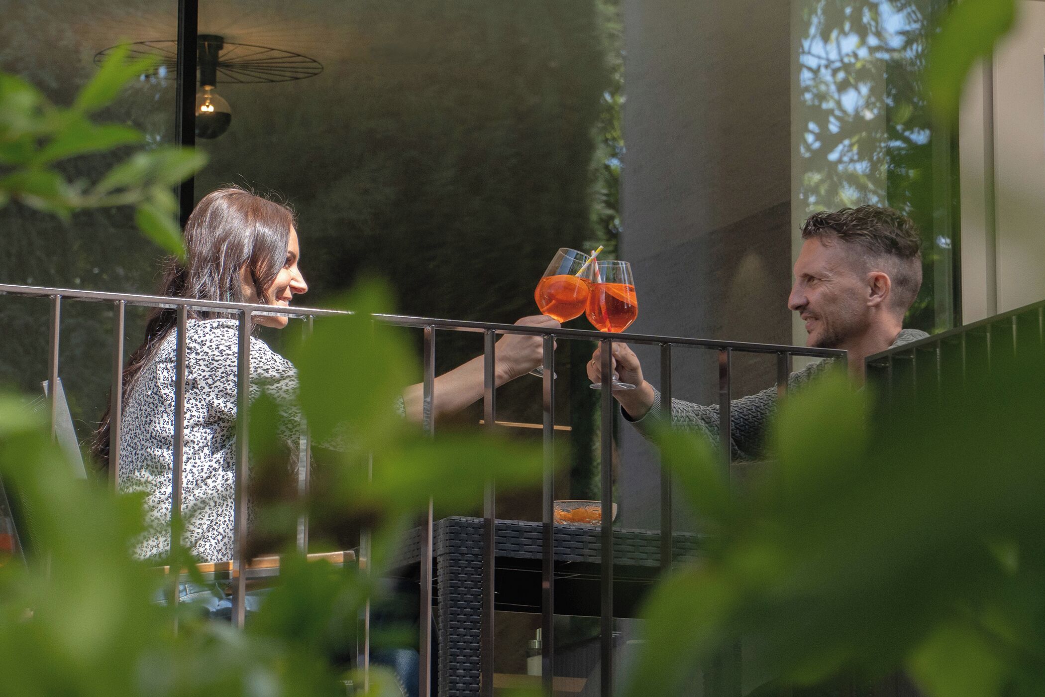 Dos personas brindando con bebidas en un balcón con vistas a la vegetación.
