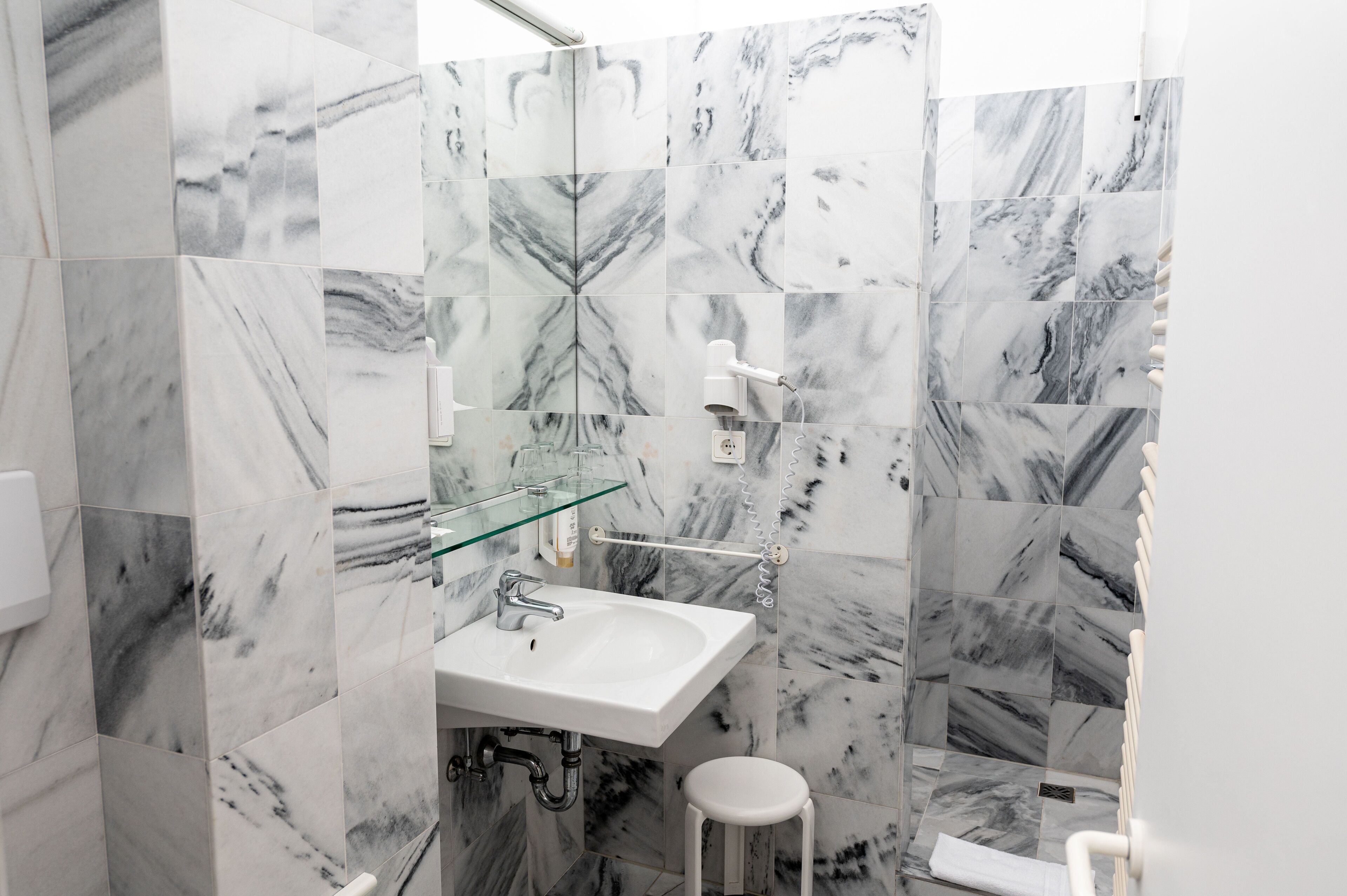 Un baño con lavamanos blanco, cabina de ducha con mampara de vidrio y baldosas con diseño de mármol.