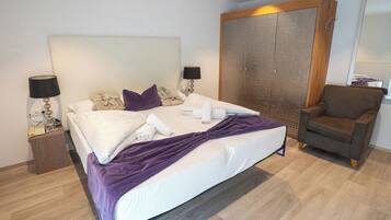 A neatly made bed with white linens and a purple throw blanket, a wooden wardrobe, a brown armchair, and two bedside lamps.