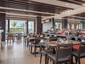 A restaurant with multiple dining tables and chairs, a buffet area with various food items, and large windows offering a view of palm trees. - The District Boracay (Boracay Island)