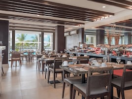 Un restaurante con varias mesas y sillas, una zona de bufé con diversas opciones de comida y amplias ventanas que ofrecen vistas a palmeras.