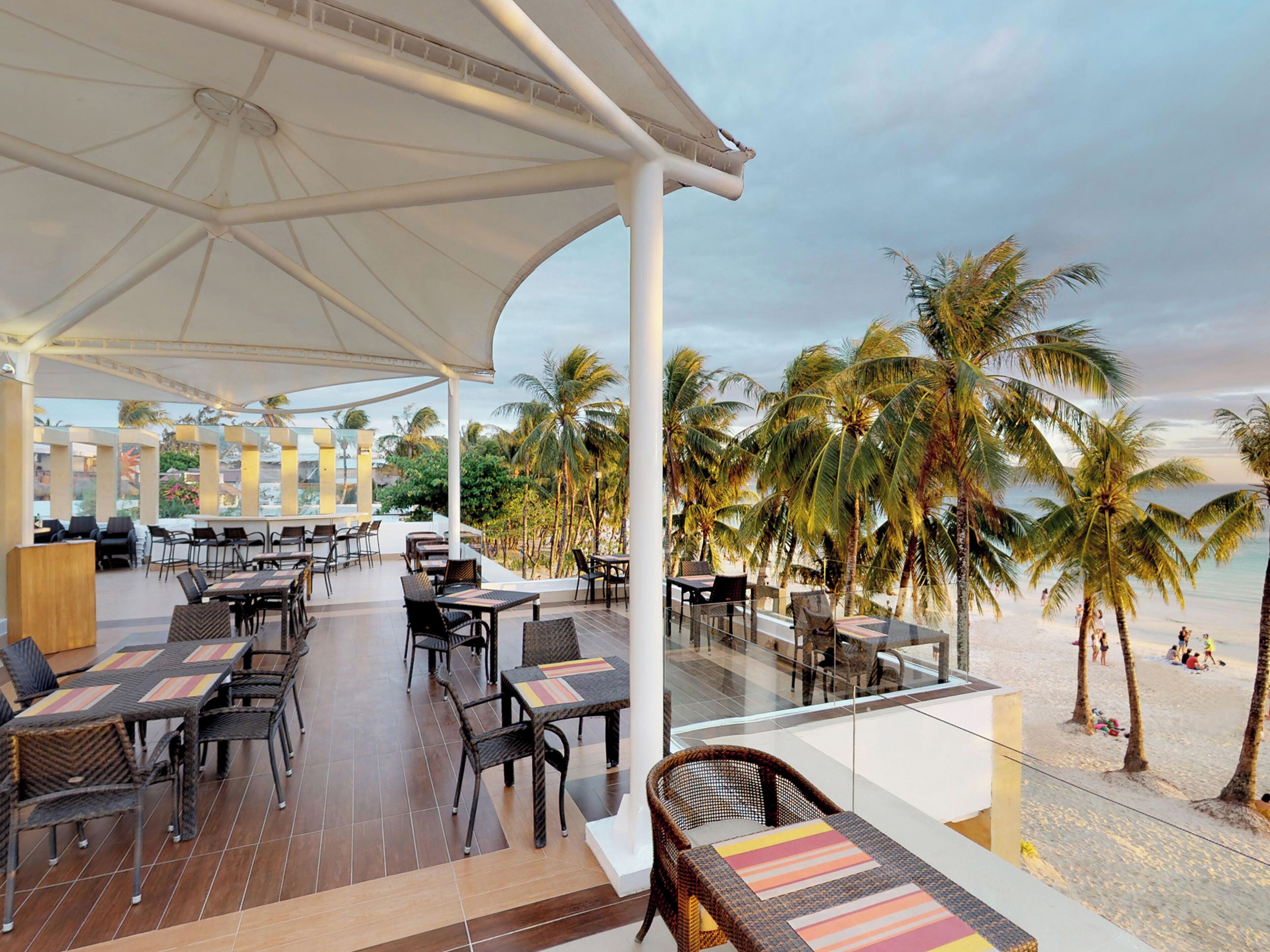 An outdoor dining area with tables and chairs, overlooking a beach with palm trees.