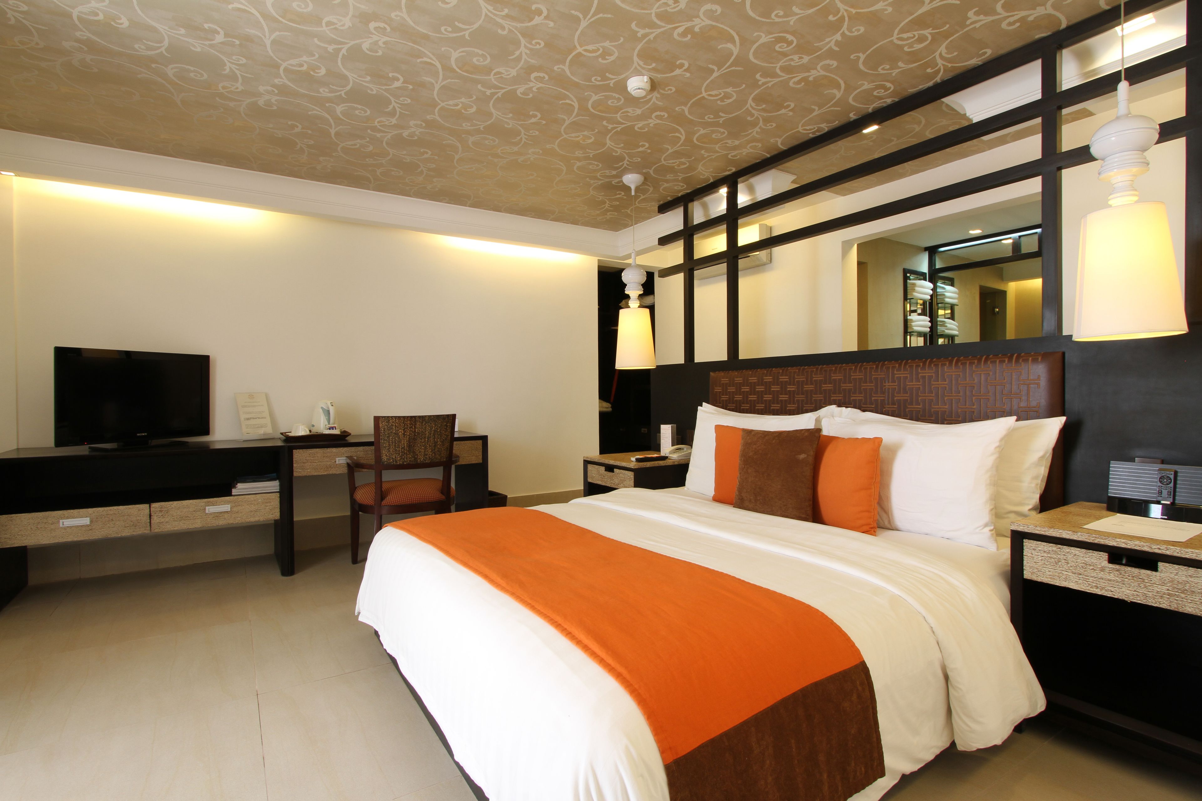 Habitación de hotel con una cama grande, un escritorio con televisor, una silla y una mesita de noche con lámpara.