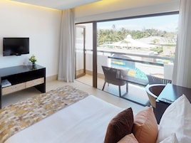 A hotel room with a bed, a TV, a desk, and a balcony with a view of a resort.
