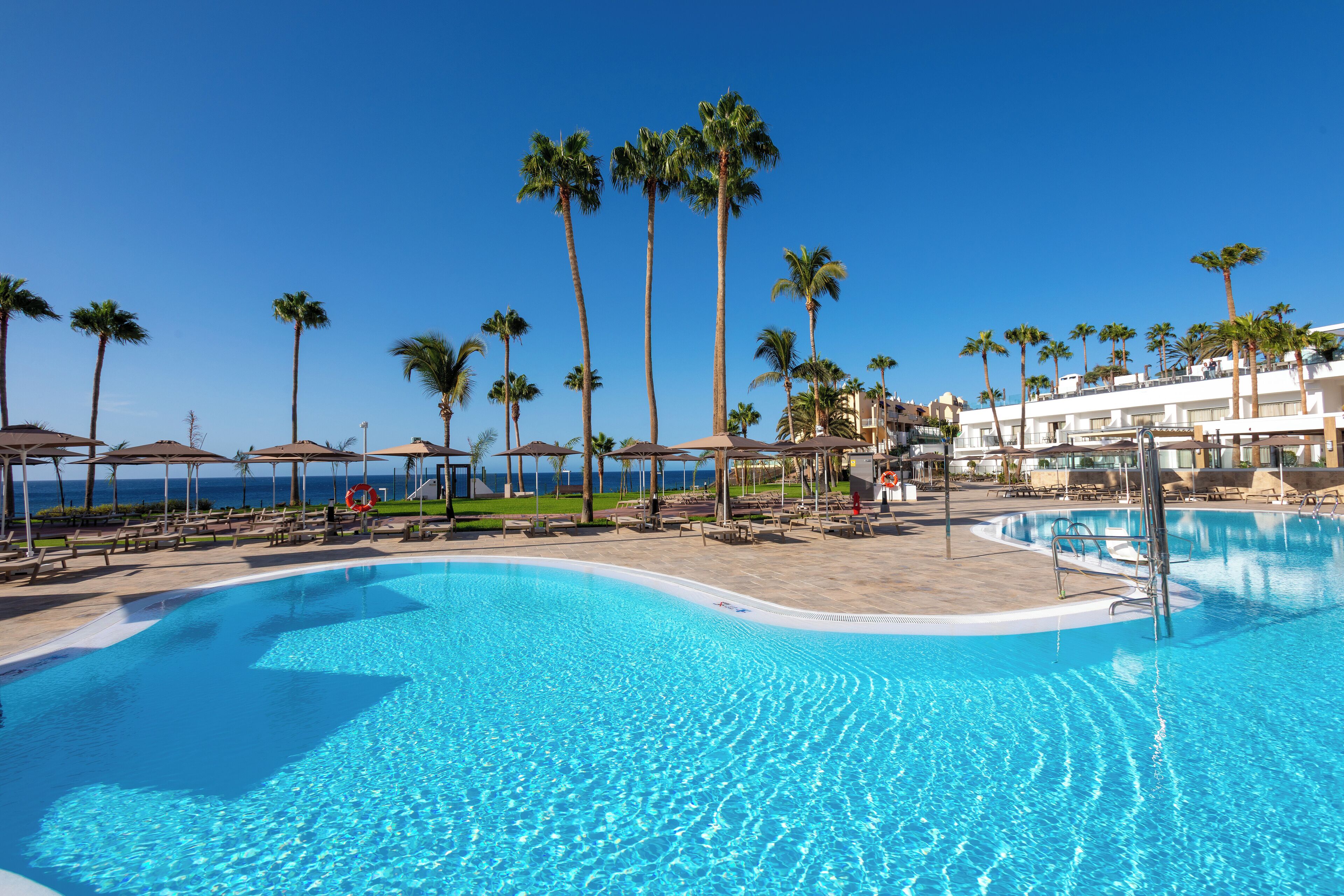 A clear blue pool with palm trees and sun loungers, surrounded by a building with a view of the ocean.